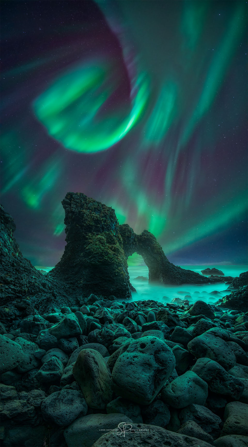 A green aurora is pictured above and beyond a dark rocky arch.
Faint stars dot the background.
Please see the explanation for more detailed information.