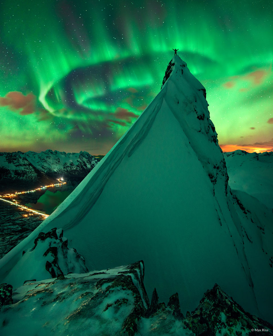 A person stands on a steep snow-covered hill with their 
arms raised. In the distance green aurora are visible. Past that
stars are visible.
Please see the explanation for more detailed information.