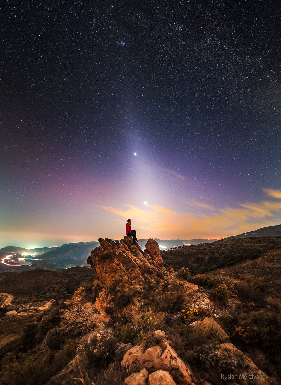 A person is seen sitting on a rock under an unusual sky. In the 
sky above is light diffuse band extending down to the horizon that 
goes through two bright dots, Jupiter and Venus. The Pleiades 
star cluster is visible above them.
Please see the explanation for more detailed information.