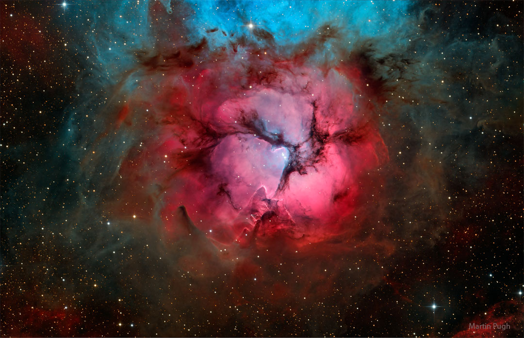 A bright red gaseous nebula is pictures with three dark
dust lanes meeting in the center. The top of the nebula appears
blue.
Please see the explanation for more detailed information.