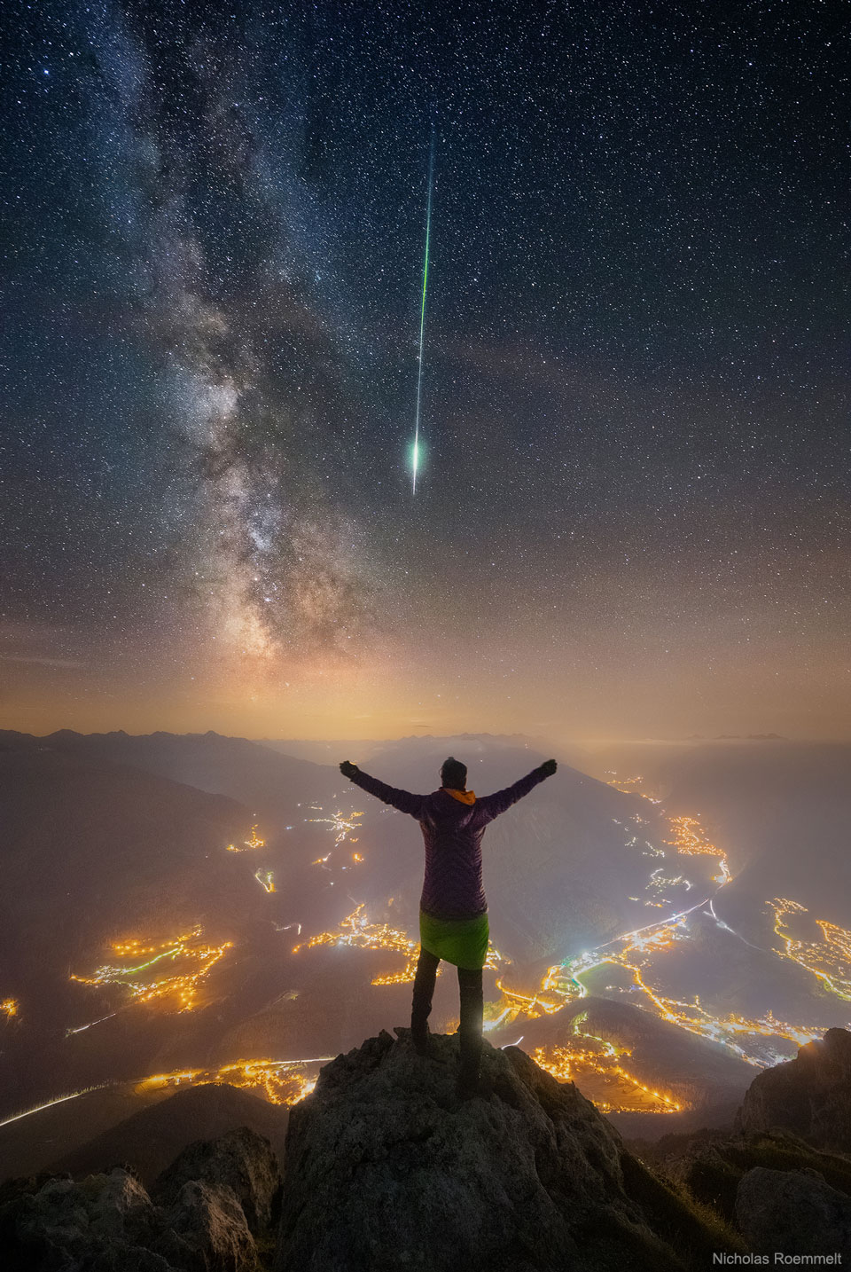 A person is seen facing away, standing on a peak. Other mountain peaks
surround them. City lights are seen in towns and along roads below. Stars
in the night sky are above. The band of the Milky Way galaxy slants down
from the upper left. A bright green meteor streak slants down from above.
Please see the explanation for more detailed information.