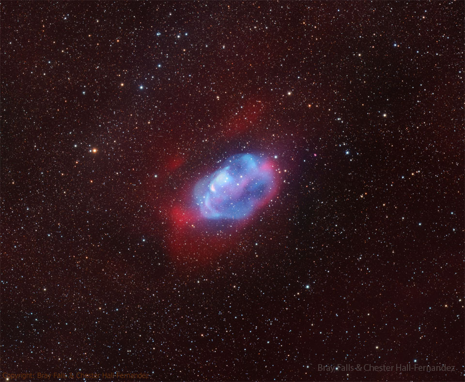 A diffuse nebula is seen against a dark starfield. The center of the 
nebula is blue and it is surrounded by a red glow.
Please see the explanation for more detailed information.