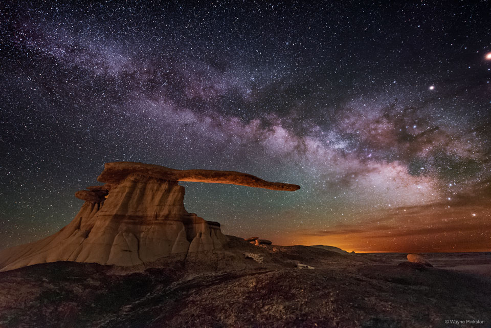 The band of the Milky Way runs across a night sky filled 
with stars. Colorful clouds are on the right horizon. A strange
rock structure appears in the image center with a base and 
an extended arm that seems to point to the colorful horizon.
Please see the explanation for more detailed information.