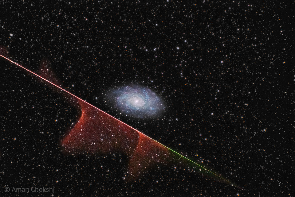 A distant spiral galaxy is seen in the image center.
A multi-colored streak runs diagonally across the image
from the upper left to the lower right. Parts of this
streak have gas near it. The background is dark field
filled with stars. 
Please see the explanation for more detailed information.