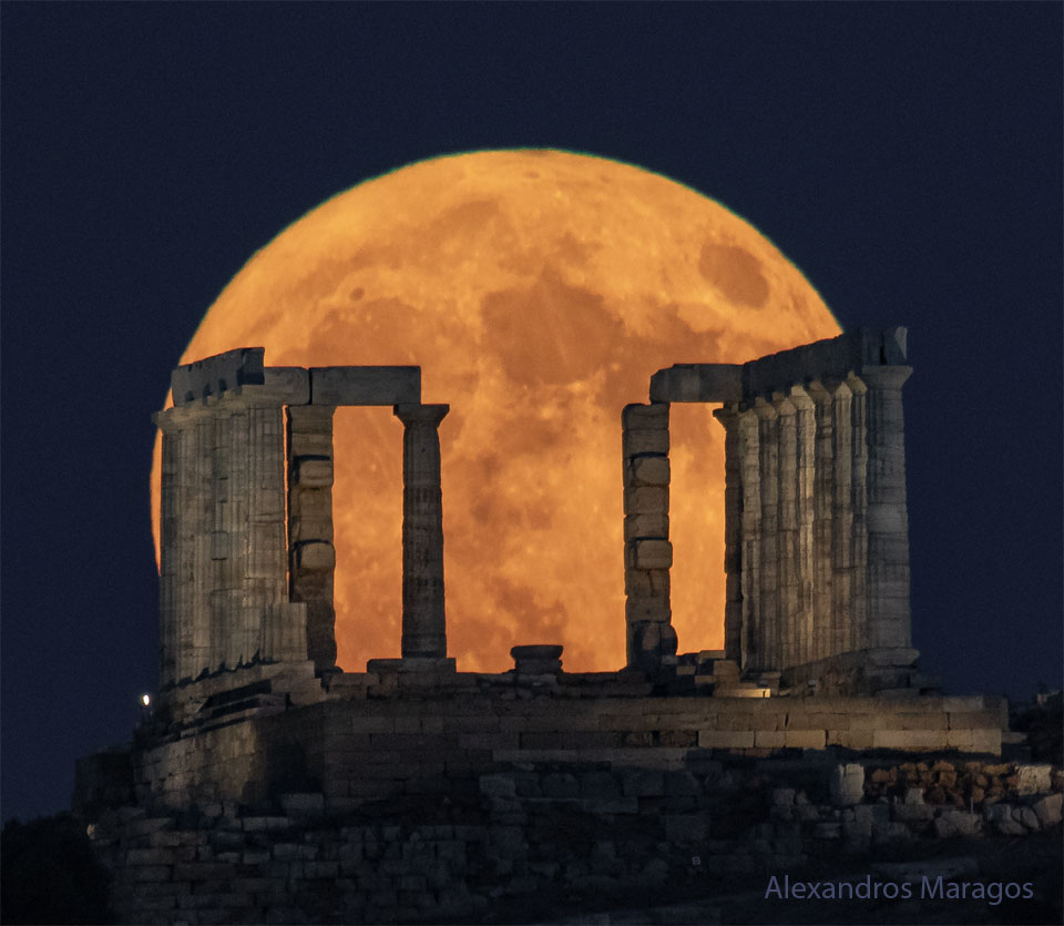 A large and orange-tinted moon is pictured rising beyond
the pillars of an ancient structure. The foreground is dark and
the night sky behind the Moon appear blue. 
Please see the explanation for more detailed information.