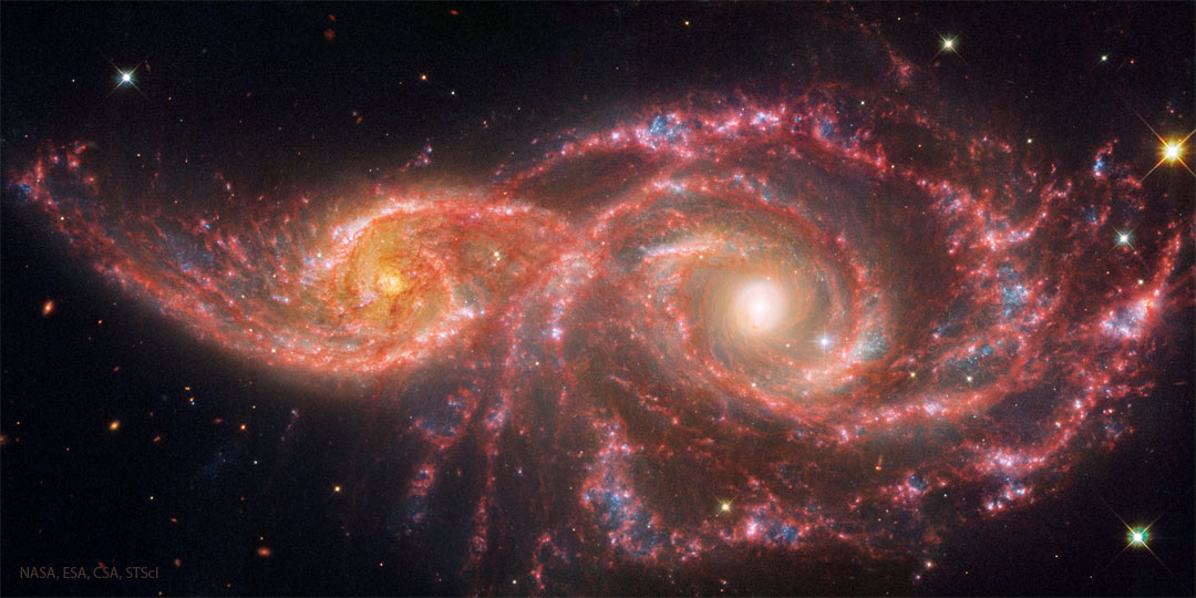 Two spiral galaxies are pictured on the left and right. 
They galaxy on the left is smaller. Both show red lanes of
dust in their spiral arms. 
Please see the explanation for more detailed information.