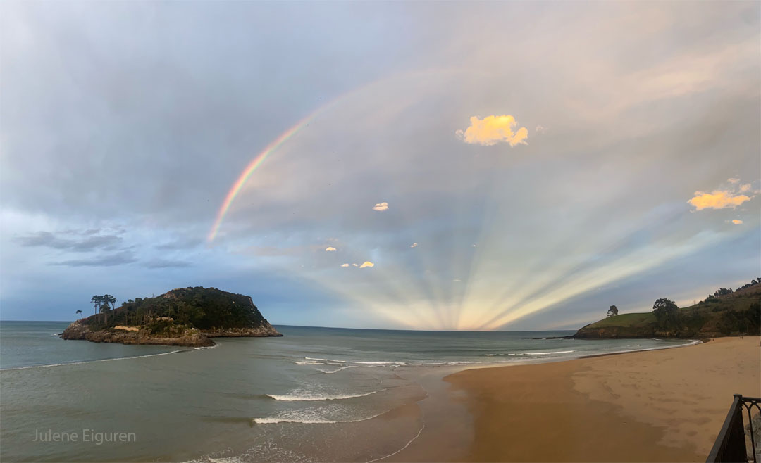 A rainbow is pictured over the sea between an island
and land. A series of
light rays appears to connect the horizon to the rainbow.
Please see the explanation for more detailed information.