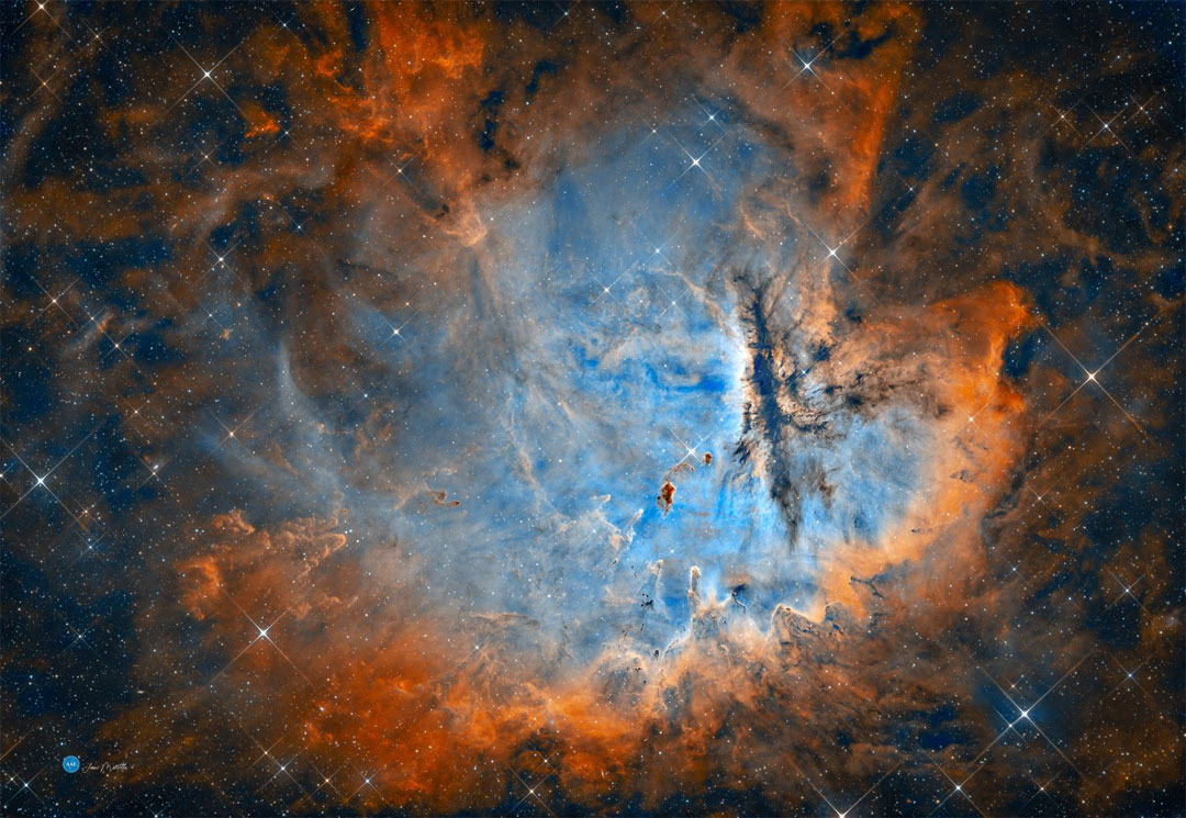 A starfield dominated by a large nebula is pictured. 
The center is blue and the perimeter is red. Many dark 
dust pillars are visible. 
Please see the explanation for more detailed information.