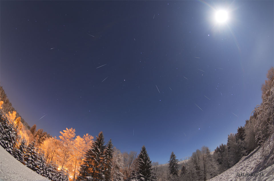 A snowy landscape is pictured below a starry sky. The very
bright Moon appears on the upper right. Many streaks are visile
that are meteors taken over the night. 
Please see the explanation for more detailed information.