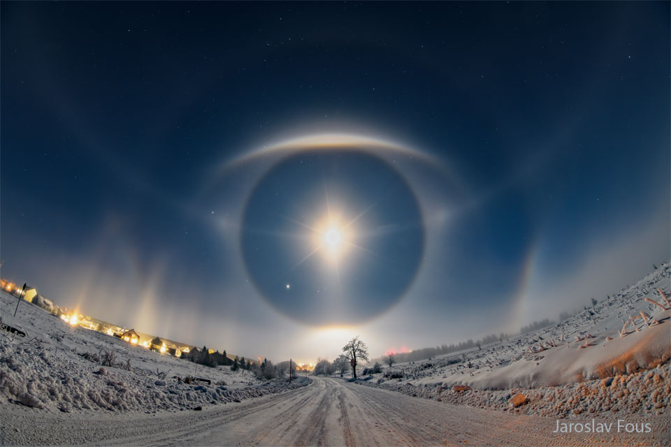 A snow covered road goes up a hill to a sky filled with
stars. Arcs and halos in the sky ahead appear similar to a giant
eye. 
Please see the explanation for more detailed information.