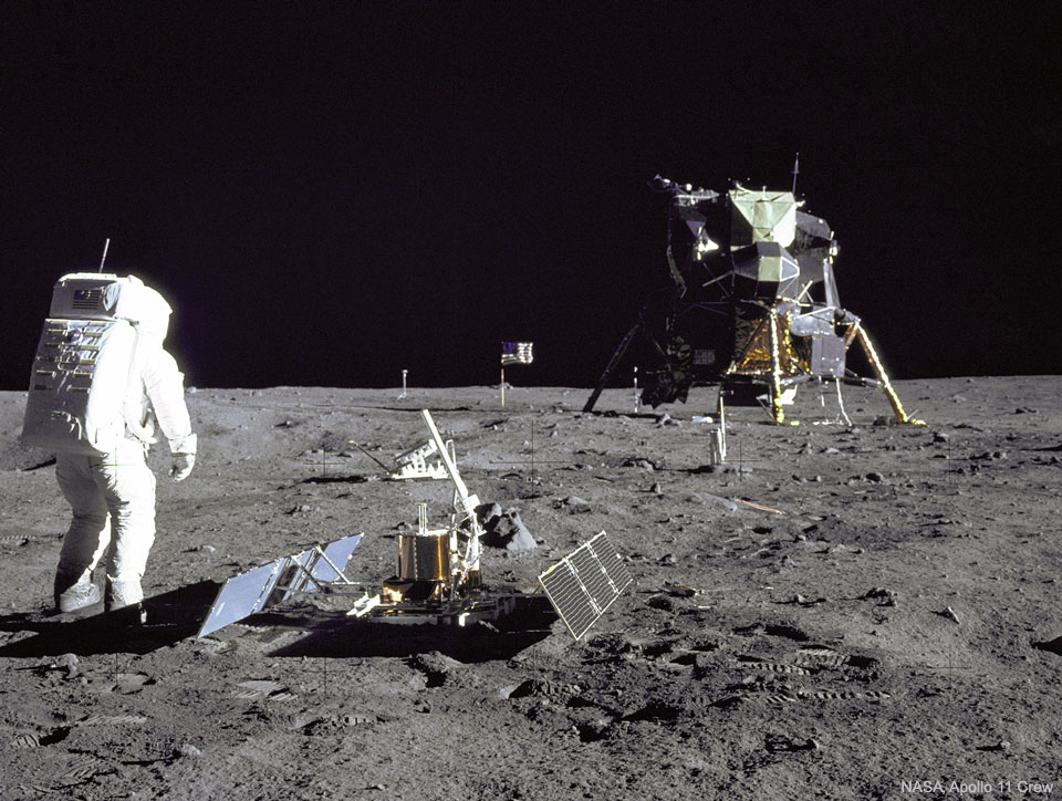 An astronaut is seen standing on the Moon next to 
an instrument with the lunar lander several meters in the 
distance. The dark of deep space covers the top of the frame. 
Please see the explanation for more detailed information.