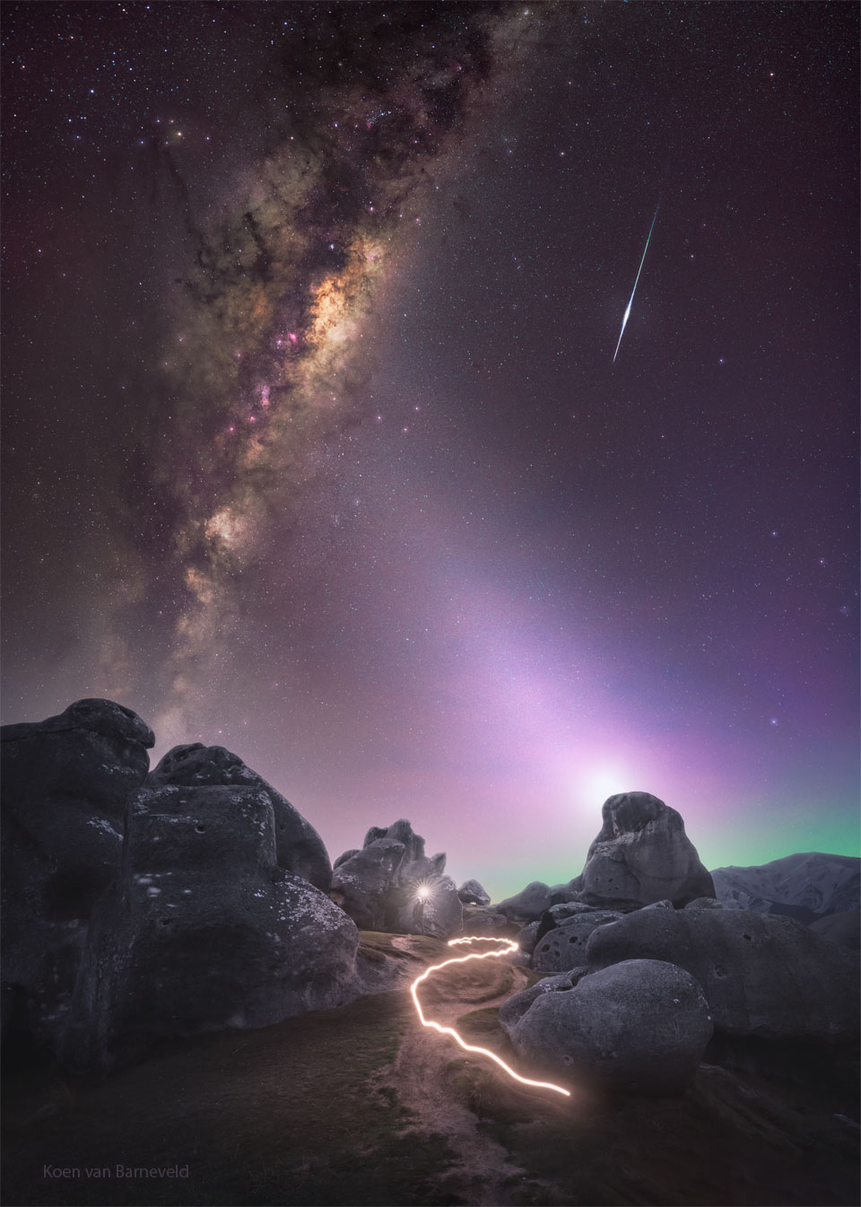 A skyscape is shown over a rocky landscape. In the starry sky
are the central band of our Milky Way Galaxy on the left, a 
meteor trail on the right, the dim band of zodiacal light 
in the center, and the photographer holding a light just below
the center. The path of the light is shown as a bright streak
in the bottom part of the frame. 
Please see the explanation for more detailed information.