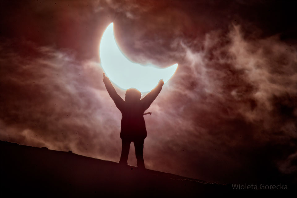 A a cloudy sky appears with an overall pink - red hue.
The Sun appears partially eclipsed over a slanting hill. 
A person on the hill has their arms raised and appears to 
be holding up the partially eclipsed Sun. 
Please see the explanation for more detailed information.