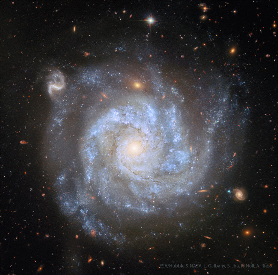 A large spiral galaxy appears with stars in the 
foreground and smaller galaxies far in the background. The
picturesque spiral has dark dust lanes between blue arms.
Please see the explanation for more detailed information.
