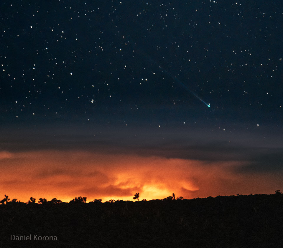 A starfield is seen above a horizon and an
orange sunset. In the starfield, near the horizon,
is a comet with a green head and long tail.
Please see the explanation for more detailed information.