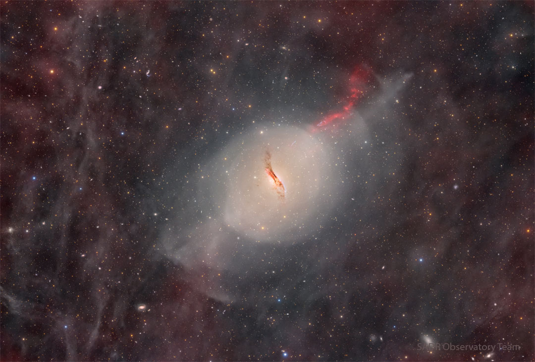 A starfield glows faintly with red glowing gas and dust.
In the center there is some sort of unusual galaxy with a red jet
emanating toward the upper right and a dark dust lane through
its center.  
Please see the explanation for more detailed information.
