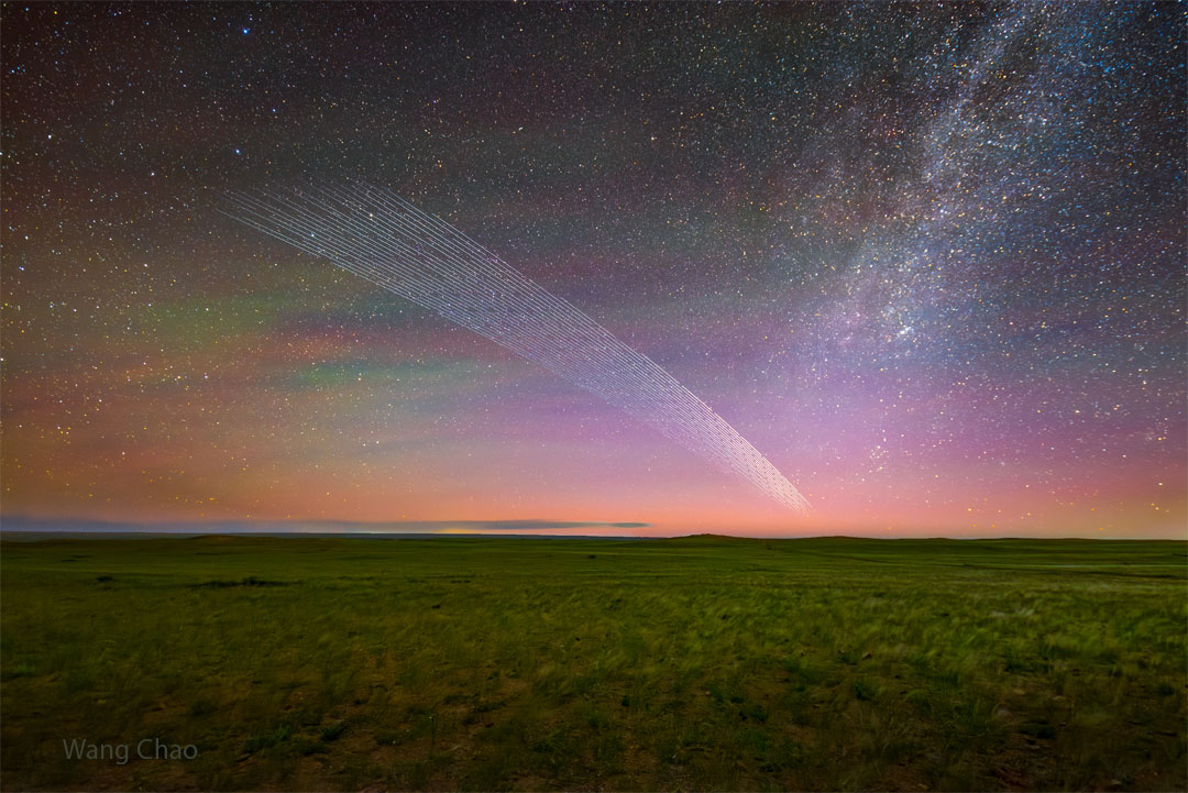 A star filled sky shows bands of green and purple sky glow. In
the foreground is a grassy field with clouds on the horizon. Most 
remarkably, a series of short streaks appear like a comet's
tail up from the horizon toward the upper left. 
Please see the explanation for more detailed information.