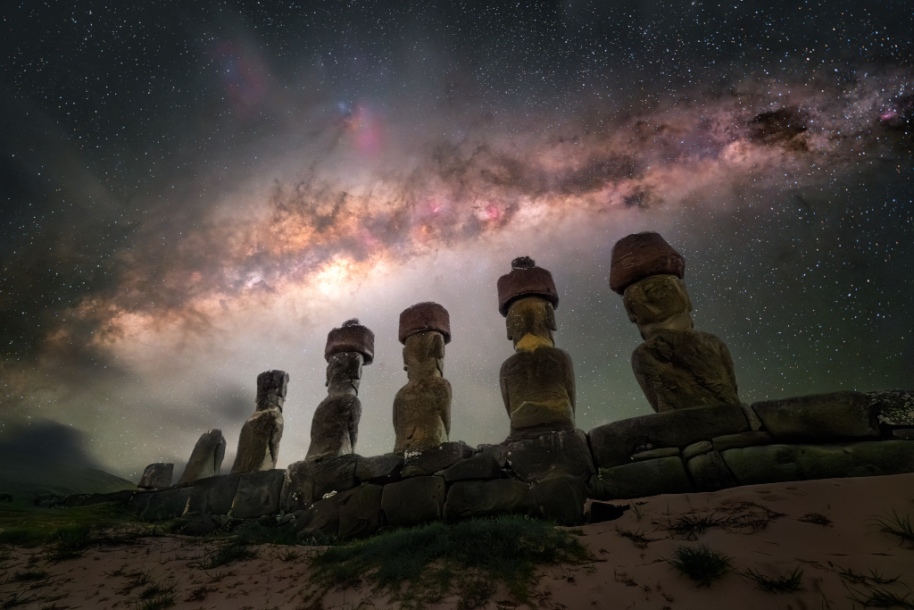 The plane of the Milky Way shines across the night sky above several 
large human statues that face away from the camera.